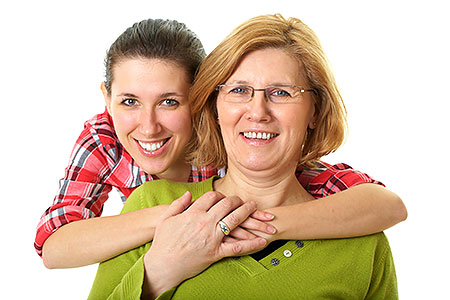 Das Foto zeigt eine Mutter, die von ihrer hinter ihr stehenden Tochter umarmt wird.